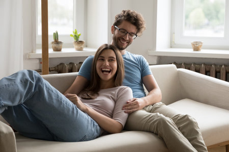 Laughing in love. Carefree happy millennial married couple share cheerful moment of rest on sofa smiling husband cuddle tickle giggling wife spend leisure time together with warmth playful affectionの写真素材
