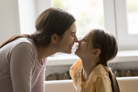 Sweet bonding ritual. Joyful little child daughter loving young mother playing together gently touching noses with soft smiles share emotional connection playful affection love sitting on comfy couchの写真素材