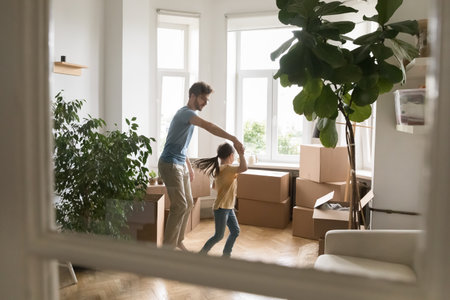 First dance in new home. Loving millennial father dancing waltz with little daughter around large light room of new apartment among packed boxes houseplants on relocation day. View through glass doorの写真素材