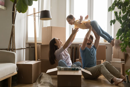 Excitement of new home. Happy family with kid flat buyers play on floor while unpacking boxes. Joyful dad hold laughing girl up in air imitate flight toward smiling mom enjoy freedom of new beginningの写真素材
