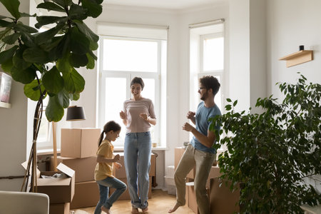 Relocation day party. Excited carefree family young couple with little daughter celebrate buying renting new flat having active fun dance jump to joyful music in living room filled with unpacked boxesの写真素材