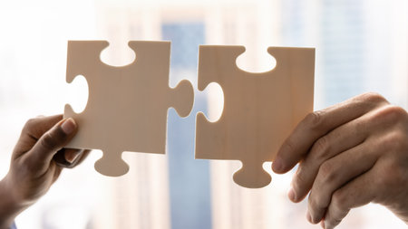 Close up view of two hands holding white pieces of jigsaw, trying to fit puzzle pieces together to...