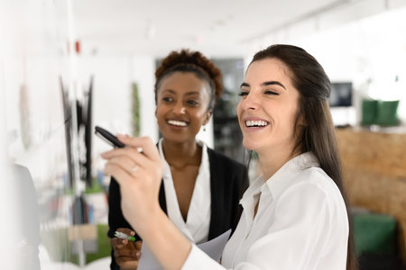 Two smiling women associates brainstorm ideas and write them on white board, close up. Teammates collaboratively planning project or strategy, share insights, visualizing data or information togetherの写真素材