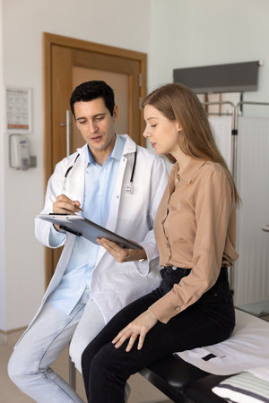 Serious medical practitioner man explaining prescription to female patient, pointing at paper document on clipboard, speaking, sitting on clinic couch, giving advice for treatment. Vertical shotの写真素材