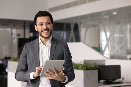 Cheerful young office employee man typing on tablet, standing in open office space alone, looking away, smiling, laughing, thinking on work tasks, ideas for business projectの写真素材