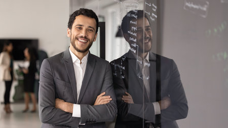 Happy young Middle Eastern business product manager man standing close to training glass board with handwritten notes, keeping hands crossed in confidence gesture, looking at camera, smilingの写真素材
