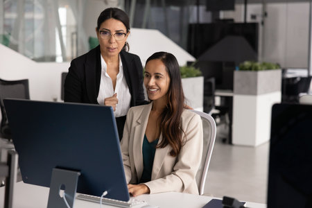 Senior businesswoman standing by happy young New Zealander professional working at computer, looking at monitor, speaking, asking for help, giving advice. Diverse colleagues meeting at pc for teamworkの写真素材