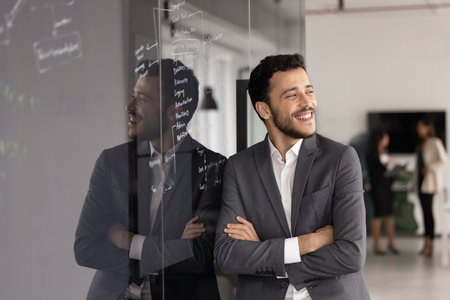 Happy young business project manager man standing at glass board in training boardroom, preparing presentation for meeting, keeping arms folded, looking away, smiling, laughingの写真素材