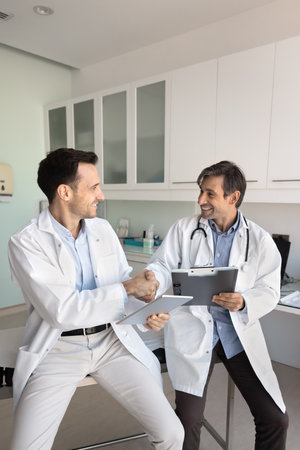 Healthcare business and partnership. Two men in white coats hold tablet clipboard shake hands in hospital office. Pharma sales agent and doctor make deal agree to collaborate on new treatment programの写真素材