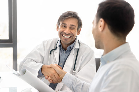 Successful job interview in medical clinic. Happy friendly chief doctor welcoming male job candidate with handshake confirm hiring greet new employee during onboarding hold folder with work contractの写真素材