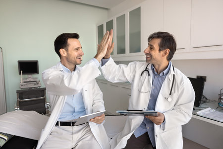 Teamwork and success in healthcare. Two smiling male doctors from diverse specialities hold digital...
