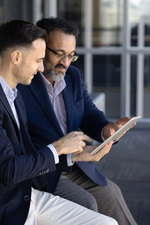 Two multiethnic businessmen in suits collaborate in office, reviewing project details on digital tablet, discussing ideas and planning next strategic steps for business, engaged in productive teamworkの写真素材