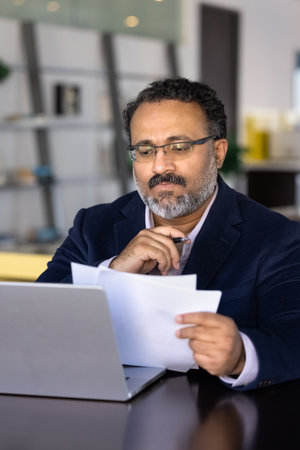 Focused businessman carefully reading printed documents while sitting at desk with open laptop, engaged in thoughtful decision-making, reviewing contract terms and conditions, learn agreement or dataの写真素材