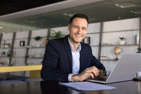 Portrait of cheerful businessman sit at desk in contemporary office, smiling at camera, working on laptop. Confidence, productive workflow using modern tech, remote collaboration, business opportunityの写真素材