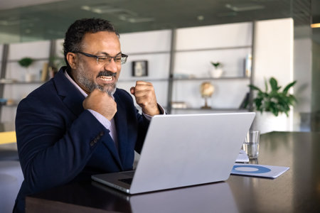 Mature 45s Indian businessman in suit sit at desk, looking at laptop screen with clenched fists and excited expression, celebrate professional success or positive outcome in work. Achievement, triumphの写真素材