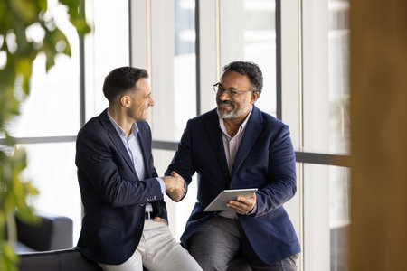 Older Indian businessman holds tablet shaking hands with Hispanic male partner or colleague met in...