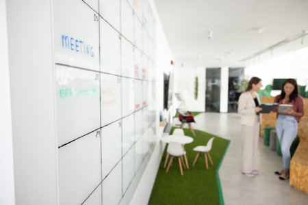 Modern office environment with large wall-mounted whiteboard calendar displaying scheduled tasks, like meeting, brainstorm, budget planning, two women professionals engaged in discussion in backgroundの写真素材
