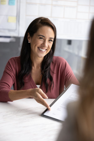 Smiling professional Latina woman presenting document to client, engaged in friendly, persuasive interaction, discussing agreement terms and conditions, proposal or contract during meeting in officeの写真素材