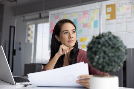 Professional woman hold analytical documents, thoughtfully looks into distance seated at desk with laptop, whiteboard with sticky notes behind her. Strategic and business planning, project managementの写真素材
