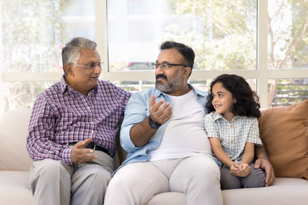 Positive Indian dad and grandad talking on couch, hugging kid girl, spending weekend, family leisure...