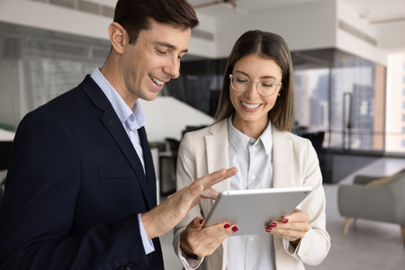 Positive coworkers using tablet for sharing ideas for project, typing, smiling, standing in office space. Businesswoman consulting colleague about electronic document, online serviceの写真素材
