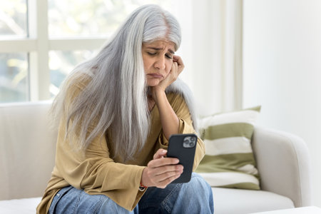 Unhappy frustrated senior Hispanic woman reading message on smartphone at home, getting bad news on...