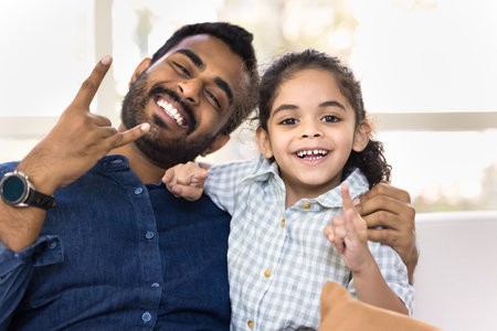 Happy joyful young Indian daddy and cute little daughter kid posing for portrait, looking at camera with toothy smile, showing rebel rock music fan hands, laughing, hugging, having funの写真素材