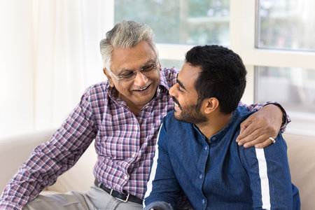 Happy Indian senior father and young adult son chatting and hugging on home sofa, enjoying family meeting, relationship, communication, friendship, sitting close, talking with trust, laughingの写真素材