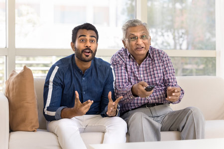 Cheerful excited young adult Indian son and older father watching sports game on TV, enjoying being...