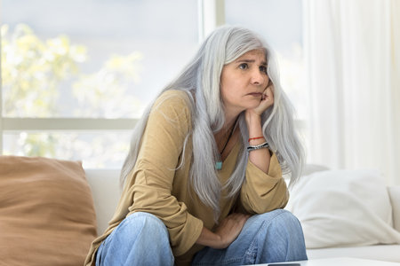 Concerned senior Latin woman sitting on home sofa, leaning chin on hand, looking away, trying to...
