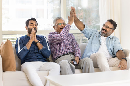 Happy joyful Indian male fans of three family generations watching sports game on TV at home,...