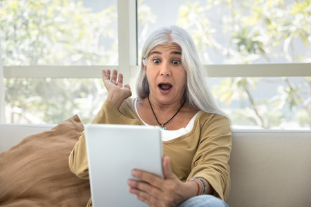 Shocked senior woman watching online content on tablet at home, looking at gadget screen with open mouth in surprise, gasping, feeling overwhelmed, making expressive hand gestureの写真素材
