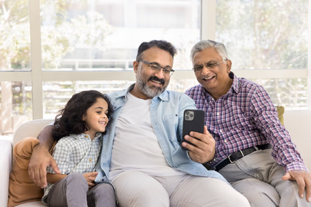 Cheerful granddad, great grandpa and grandkid girl having fun together, using mobile phone on home...