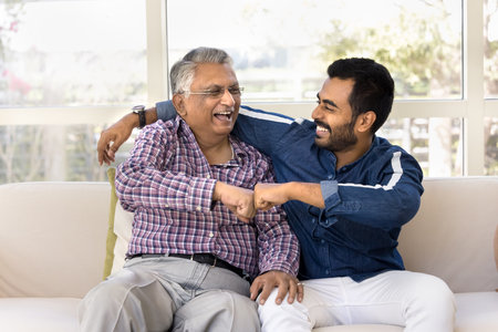Joyful excited senior Indian father and young adult son giving fist bump, celebrating successful...