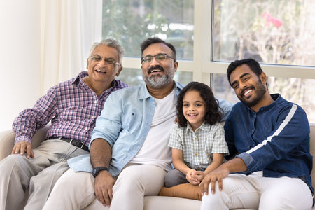 Happy adorable little Indian girl, young father, grandpa and great granddad watching TV at home, sitting on comfortable sofa close together, looking away, smiling, laughingの写真素材