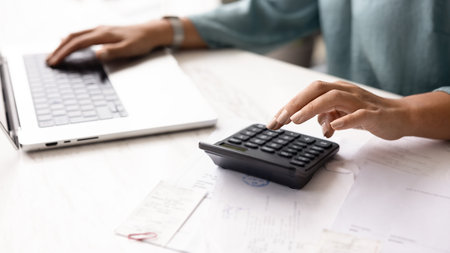 Hands of female accountant typing on calculator and laptop at office table, working on financial...