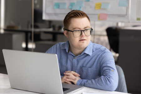 Thoughtful young professional man in glasses with Down syndrome working at laptop in office, sitting at workplace, looking away, thinking on career opportunities in inclusive companyの写真素材