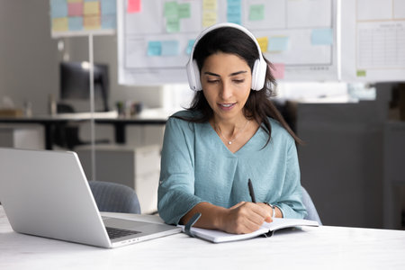 Focused young Latin businesswoman talking on video conference call, taking notes, writing in notebook during online meeting, using computer and earphones for Internet communication from officeの写真素材