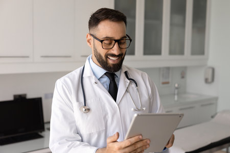Smiling Latin male doctor in eyeglasses using modern digital tablet device in examination room, check work-related e-mails, responding to patients messages through innovative platform or applicationの写真素材