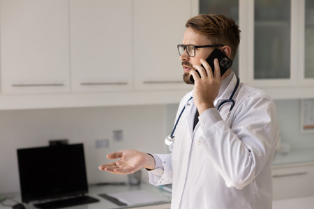 Serious male doctor standing in clinic examination room, talking on smartphone with patient, having emergency response, remote medical communication, urgent healthcare coordination, real-time supportの写真素材
