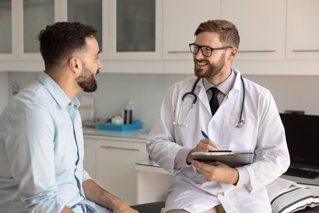 Positive doctor filling up medical card during conversation with patient in modern examination room. Latino man answers question about symptoms or conditions, passing regular health check up in clinicの写真素材