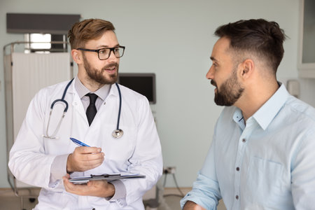 Young doctor in coat asks to man patient about symptoms, writing down in medical journal health complaints during consultation in clinic examination room. Healthcare services, professionalism, missionの写真素材