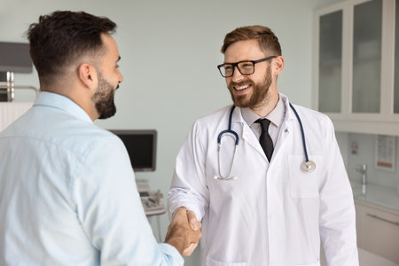 General practitioner dressed in white coat shaking hands with clinic patient, standing together in examination room, starting consultation, greet each other, provide professional healthcare servicesの写真素材