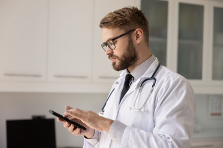Young male doctor in coat and eyeglasses stands in modern medical office with smart phone, checking medical data, communicating with colleagues, or managing patient information through healthcare appの写真素材