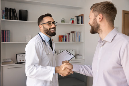 Happy Hispanic general practitioner in white coat holding clipboard, greeting male patient standing together in examination room of clinic office. Regular medical exam, check-up, healthcare servicesの写真素材