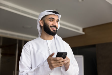Happy successful Arabian business man in white keffiyeh using mobile phone in office, holding...