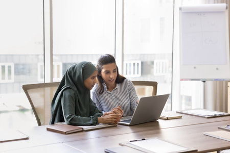 Developing sales strategy. Two diverse young female business partners Arab and Hispanic ethnicities work online use notebook at presentation room prepare financial report manage documents. Copy spaceの写真素材