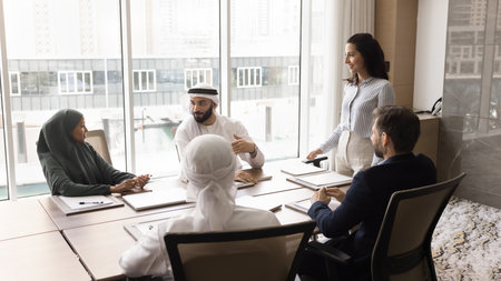 Corporate training. Young female team leader business coach stand by conference table at office lead brainstorm conversation among multiethnic company staff offer idea to discuss as teamwork projectの写真素材