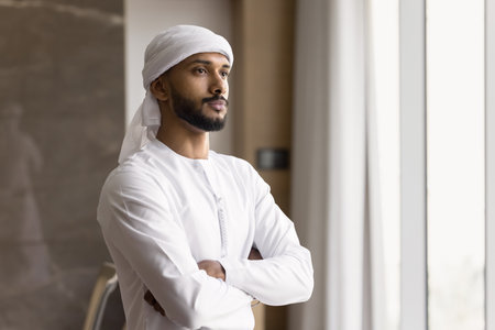 Creating ambitious plans. Thoughtful serious millennial middle eastern businessman dressed in thobe stand by window at modern office in confident pose ponder on new project estimate risks. Copy spaceの写真素材
