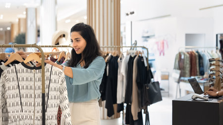 Positive young Hispanic woman viewing clothes on rack in outlet, shopping in retail fashion store. Latin sales associate girl checking prices on goods in showroomの写真素材